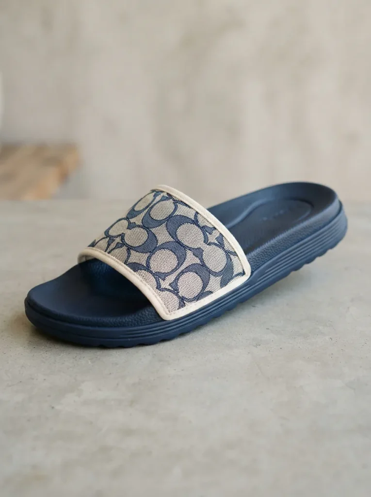 A pair of Coach Women's Udele Signature Jacquard Canvas Sport Slides displayed on a neutral background, highlighting the canvas texture and cushioned sole.